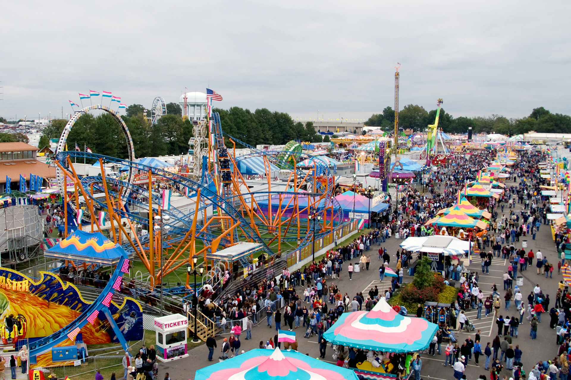 Busy fairground with colorful rides, tents, and crowds under a cloudy sky