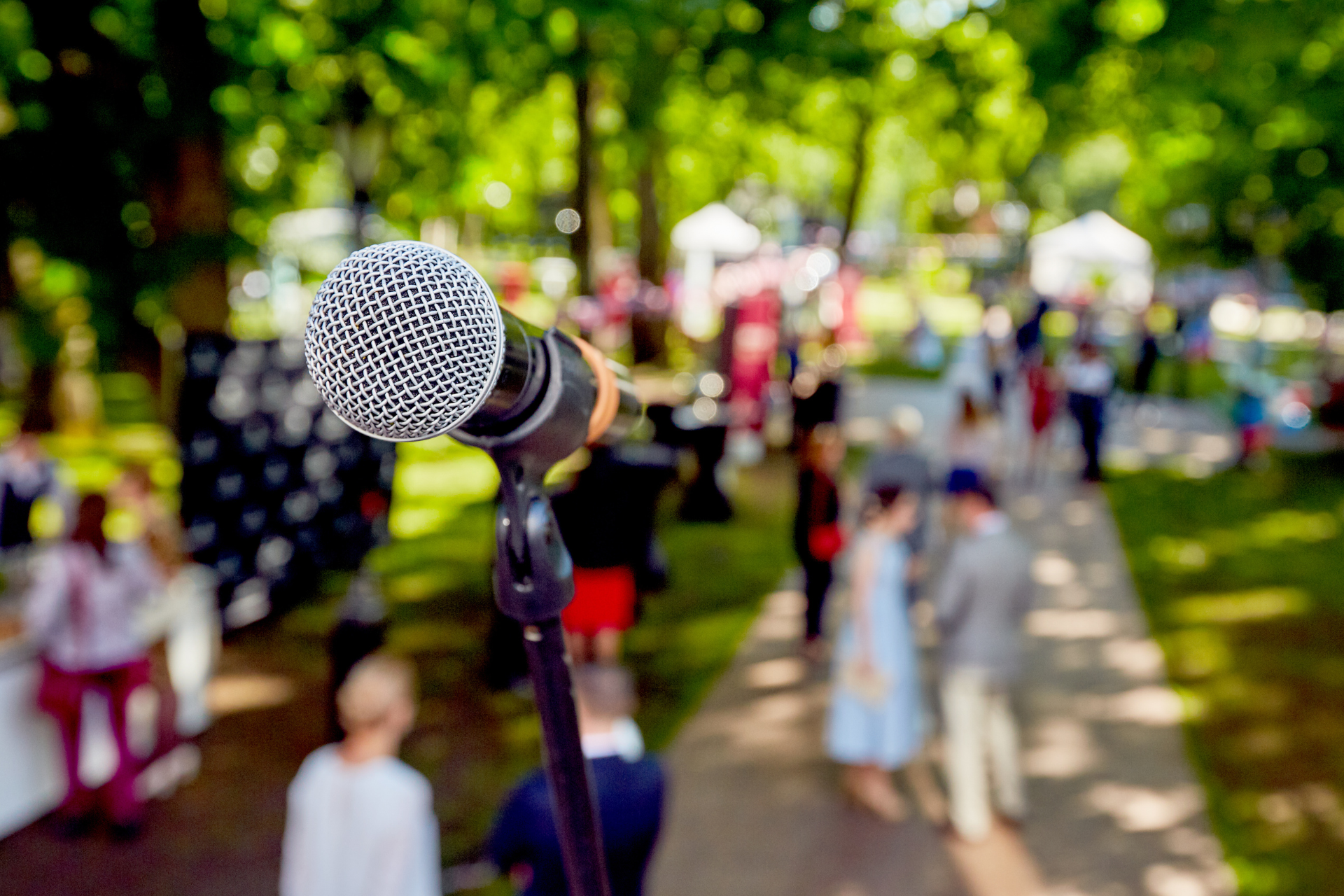Microphone on stand in foreground at an outdoor gathering with people and trees blurred in the background