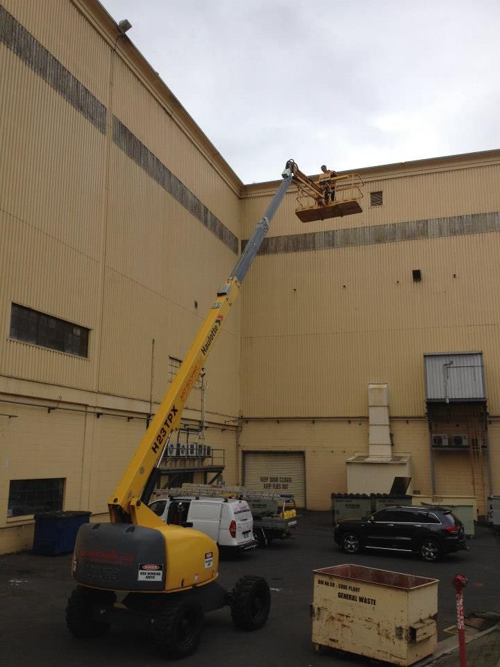 A yellow boom lift extends upward to reach the side of a tall, beige industrial building in an outdoor parking area.