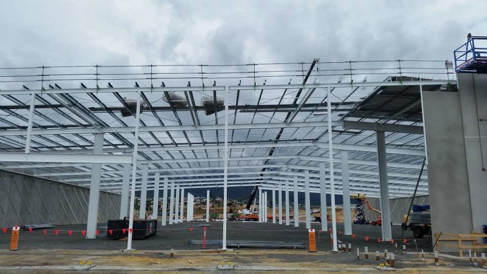 The steel frame of a large, unfinished warehouse building stands under a cloudy sky on a construction site.