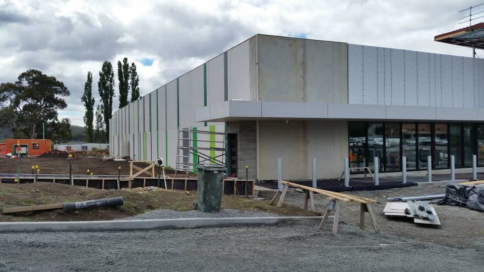 A modern, unfinished commercial building with white walls and green trim, set on a gravel construction site.
