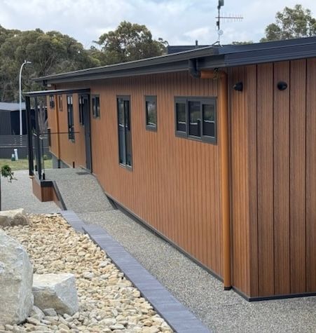 A side view of a modern building with brown vertical cladding, black window frames, and a gravel-paved accessible ramp.