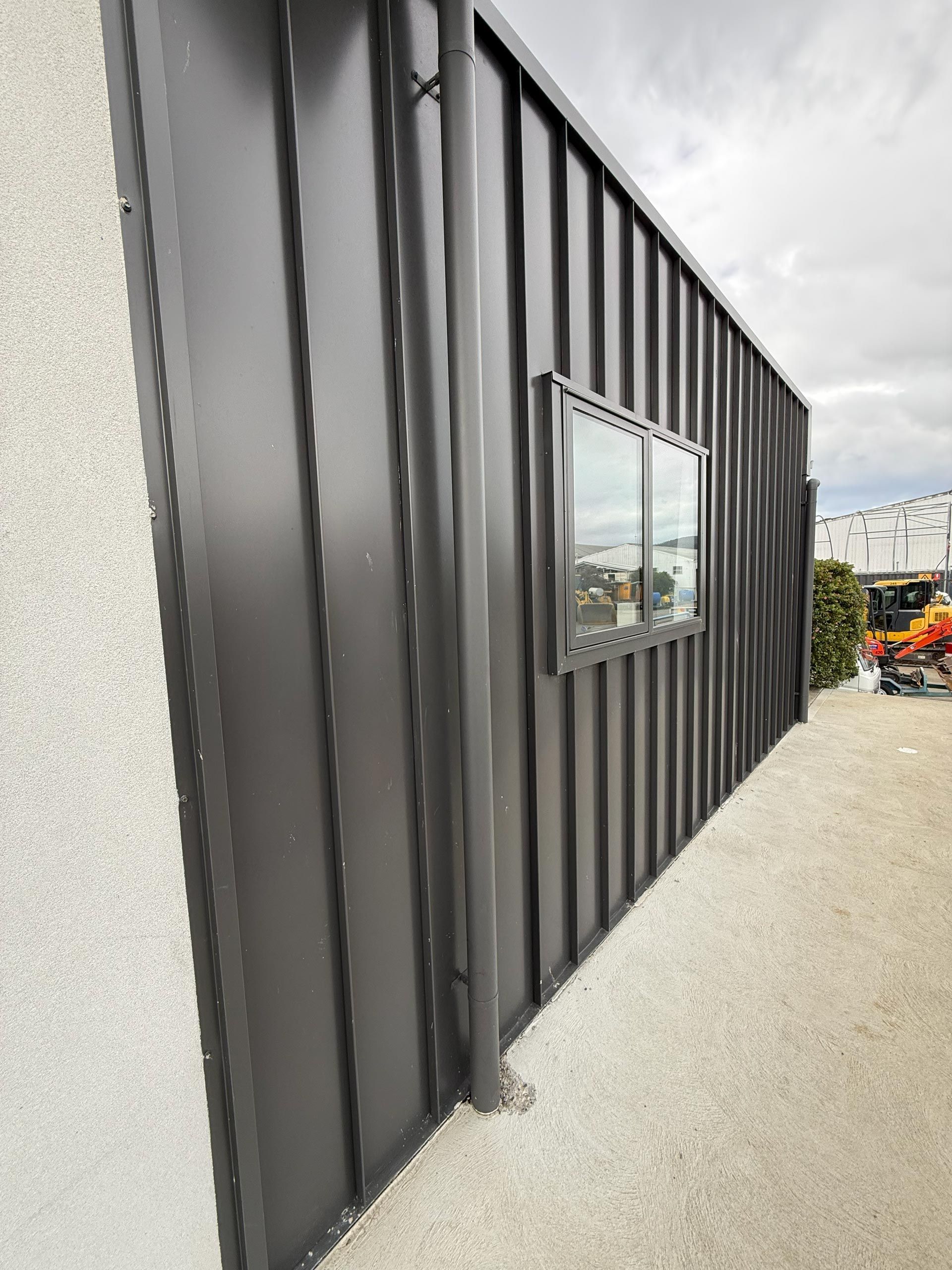 Black shipping container wall with a small window beside a gravel path outdoors