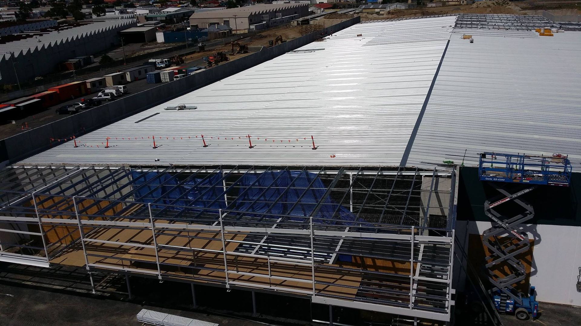 Aerial view of a commercial building under construction with a white roof, scaffolding, and a scissor lift.