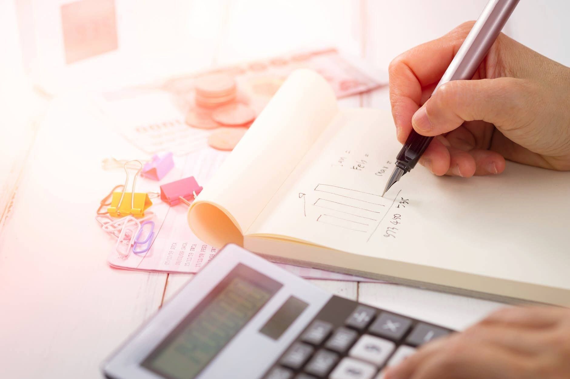 Hands writing on a financial form beside a calculator and colorful notes on a desk