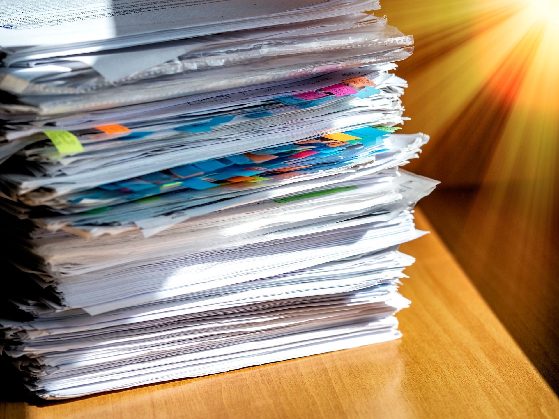 Stack of papers and folders on a wooden desk with sunlight streaming in from the right