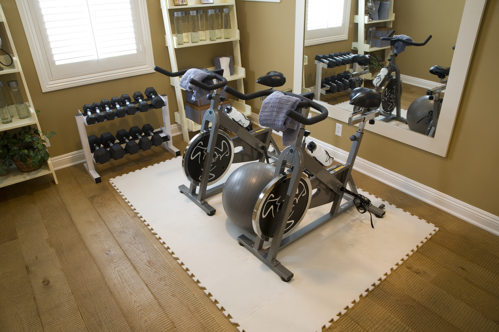 Two exercise bikes positioned on a white puzzle-mat floor in a home gym with dumbbells, a shelf, and a large wall mirror.