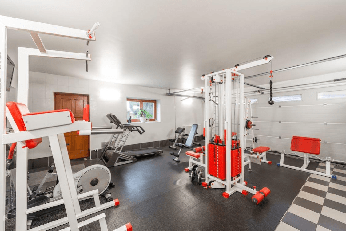 Small home gym with exercise machines, cable station, treadmill, dumbbells, and checkered floor tiles.