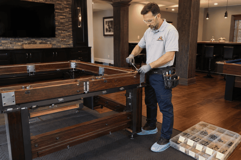 Man tightening bolts on a wooden billiards table in a living room, with tools nearby.
