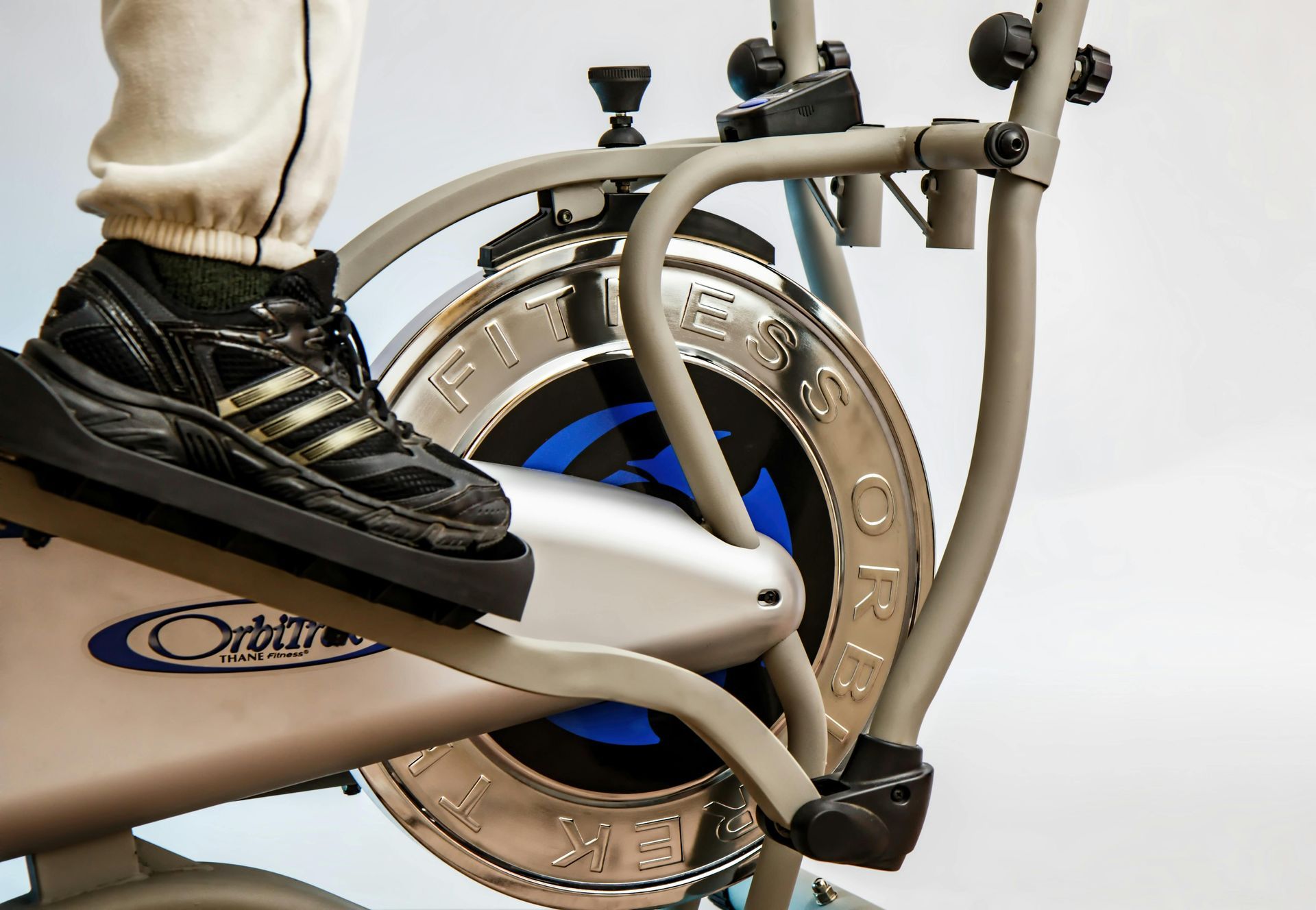 A close-up of a person's foot in a black athletic sneaker on the pedal of a grey elliptical exercise machine.