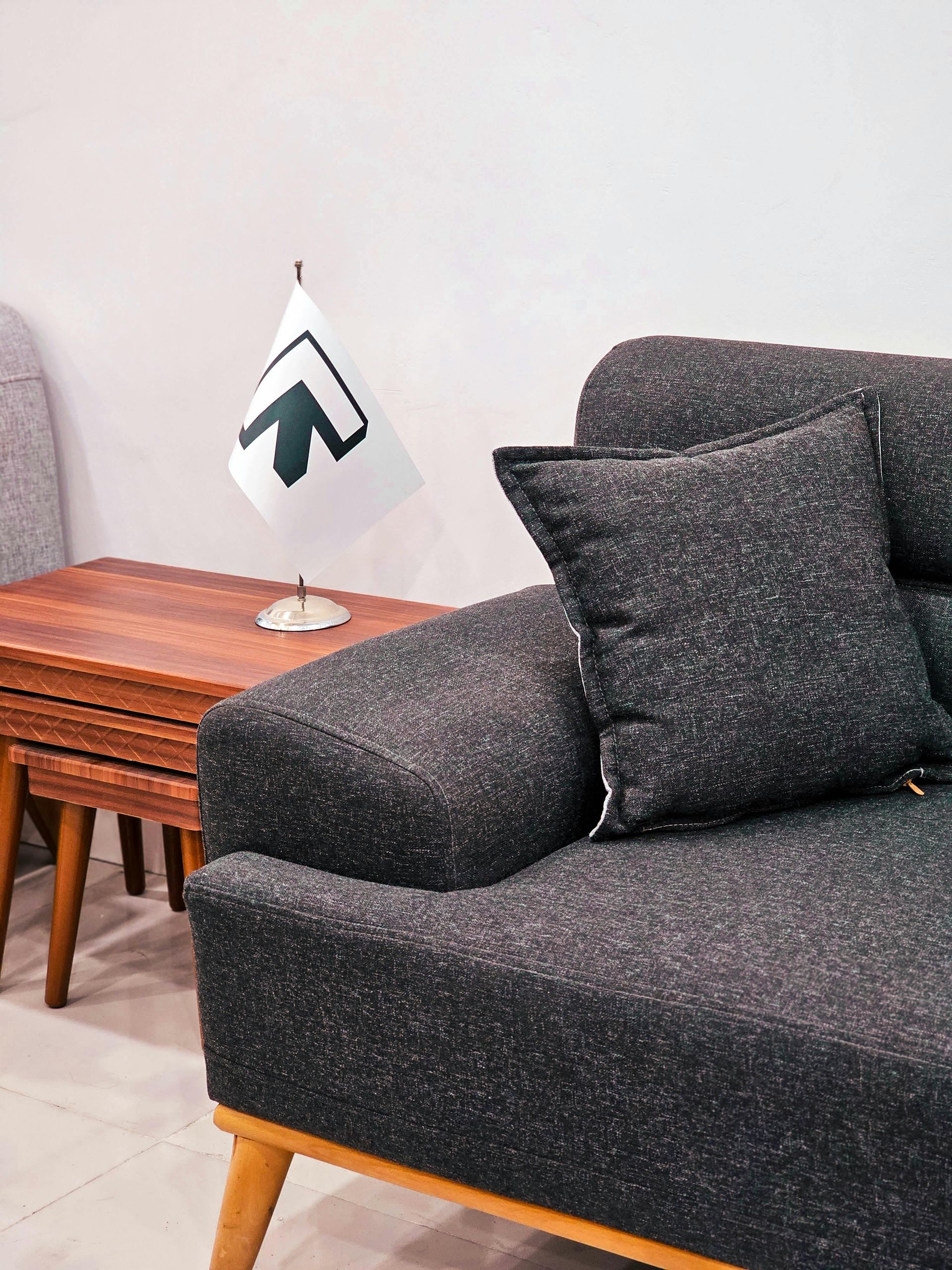 A dark, speckled fabric sofa with a matching throw pillow next to a small wooden side table holding a miniature logo flag.