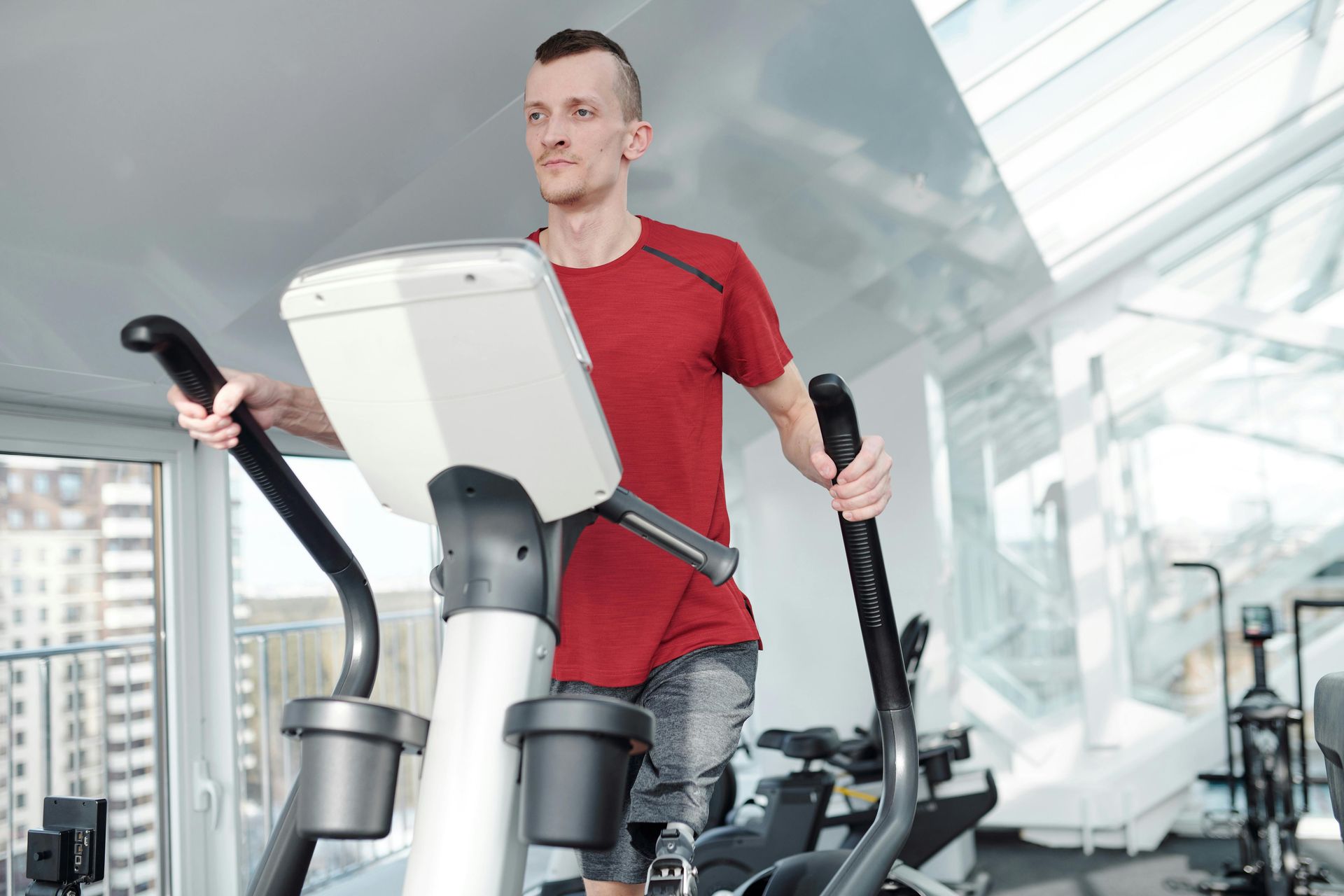 A person in a red shirt exercises on an elliptical machine in a bright, modern gym with large windows.