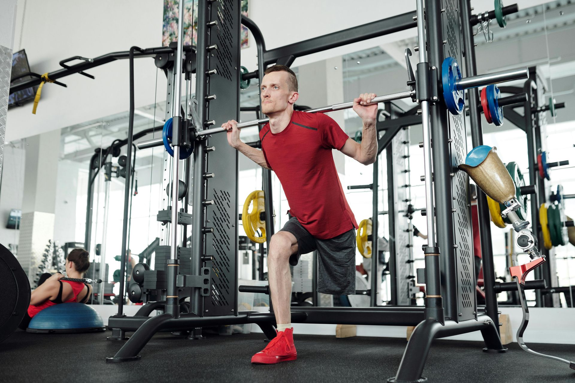 Man lunges with a barbell in a gym, wearing a red shirt and shoes, with equipment in the background