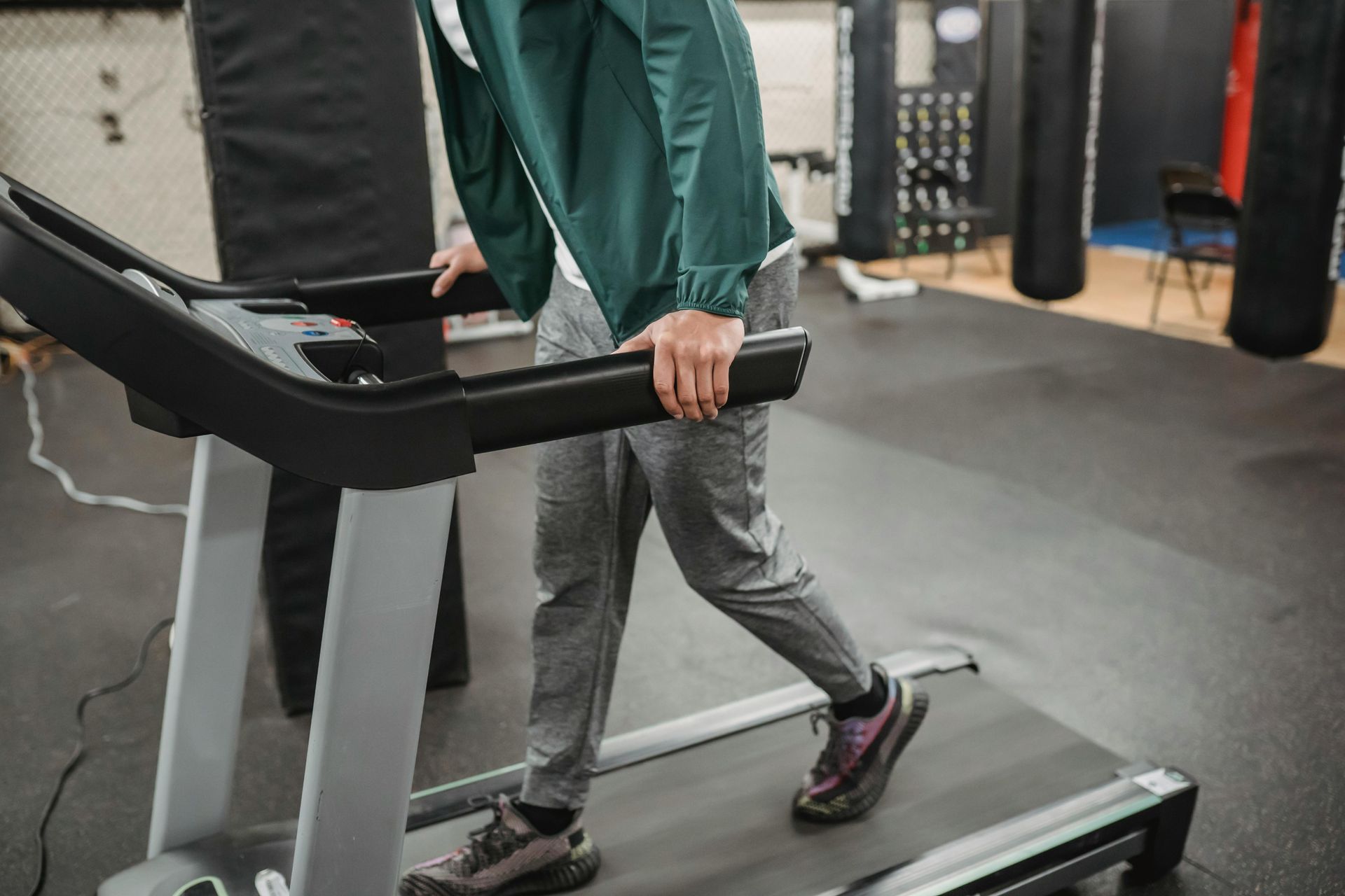 Person walking on a treadmill in a gym, wearing gray pants and a green jacket.