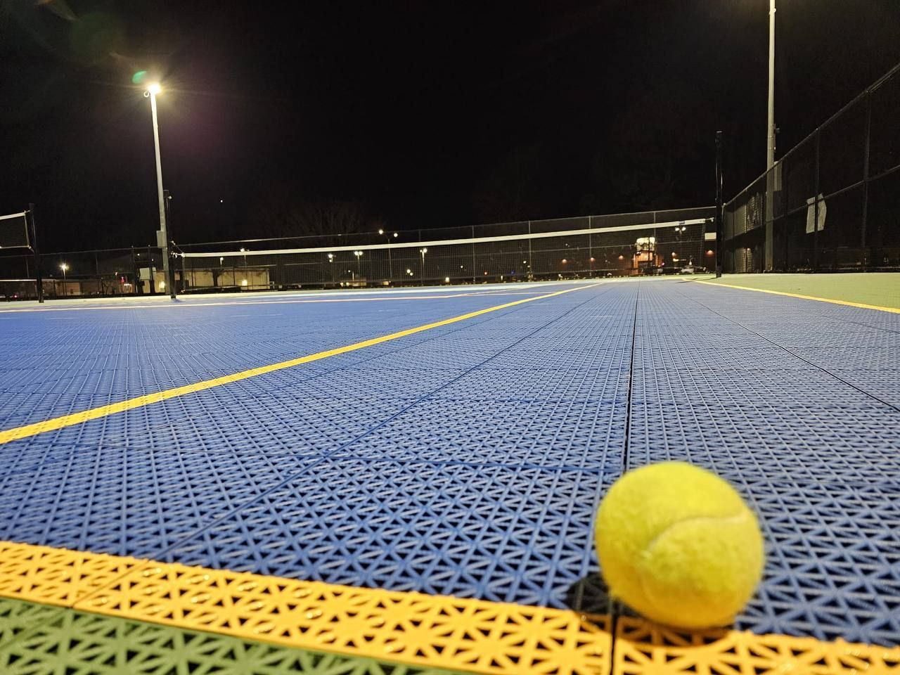 Tennis ball on a blue and yellow court at night under bright lights. Net and fencing visible.