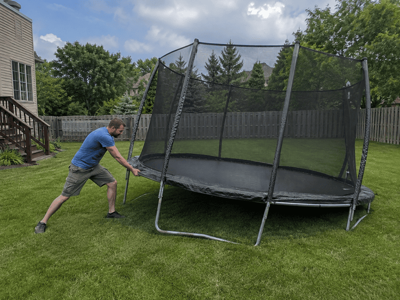 Man pushing a tilted trampoline in a grassy backyard with a fence and house nearby