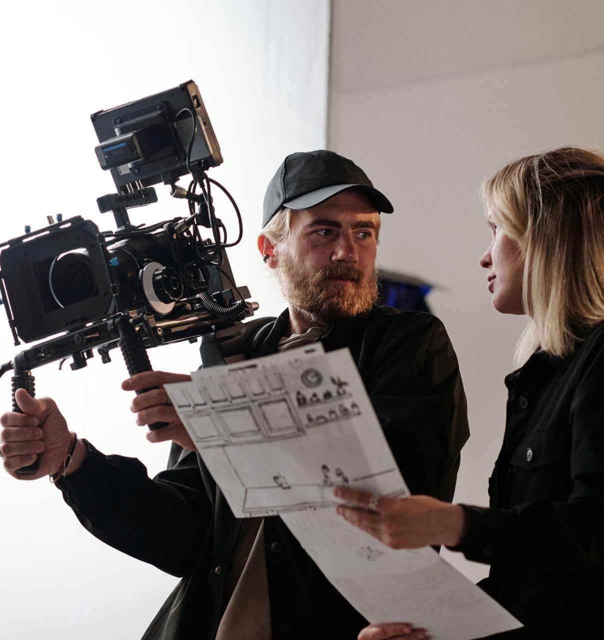 Young bearded cameraman and his assistant with documents consulting about shooting while standing in front of camera in studio using Video Branding To Increase Brand Awareness