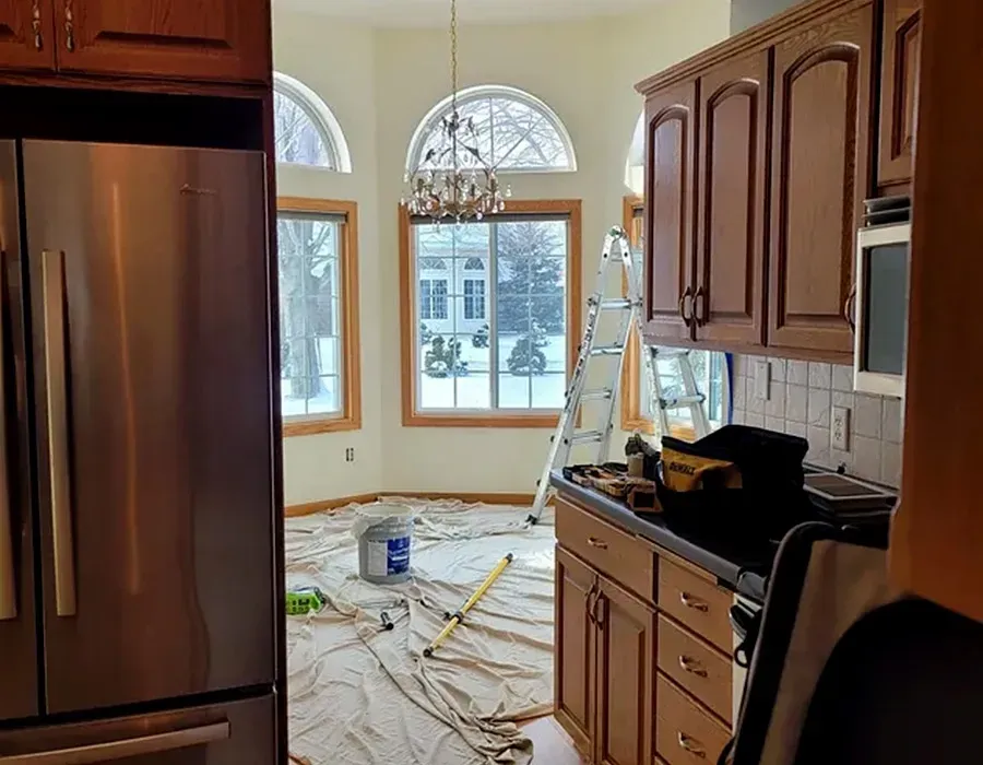 Kitchen under renovation with debris on the floor, ladder, and a windowed dining area in the background