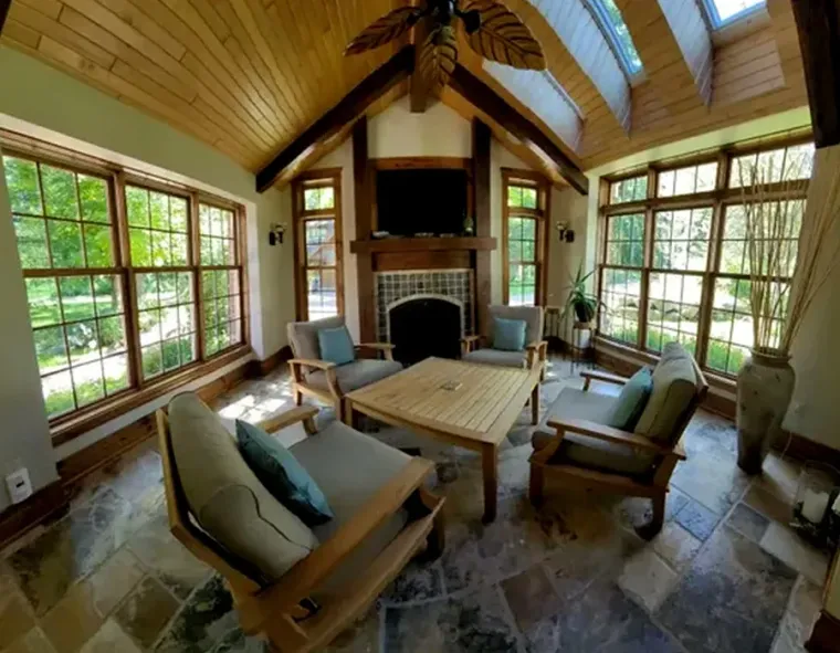 Cozy sunroom with wood beams, stone fireplace, large windows, and cushioned armchairs around a coffee table