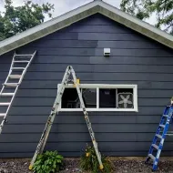 Gray house exterior with ladders set up for painting or repairs around a window and roofline