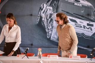 Un hombre y una mujer están sentados en una mesa frente a un automóvil que dice SM Motorsports.