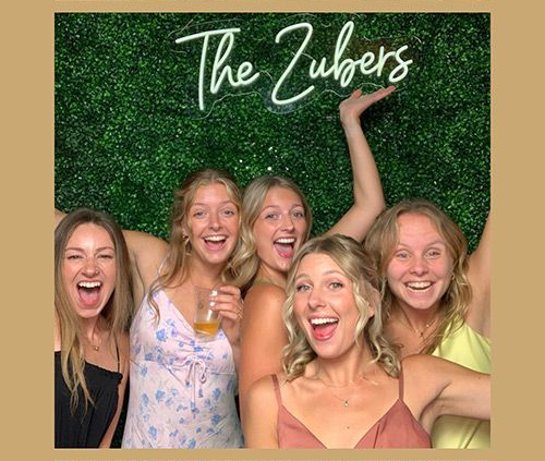 Five women smiling excitedly in a photo booth; green backdrop with "The Zubers" neon sign.
