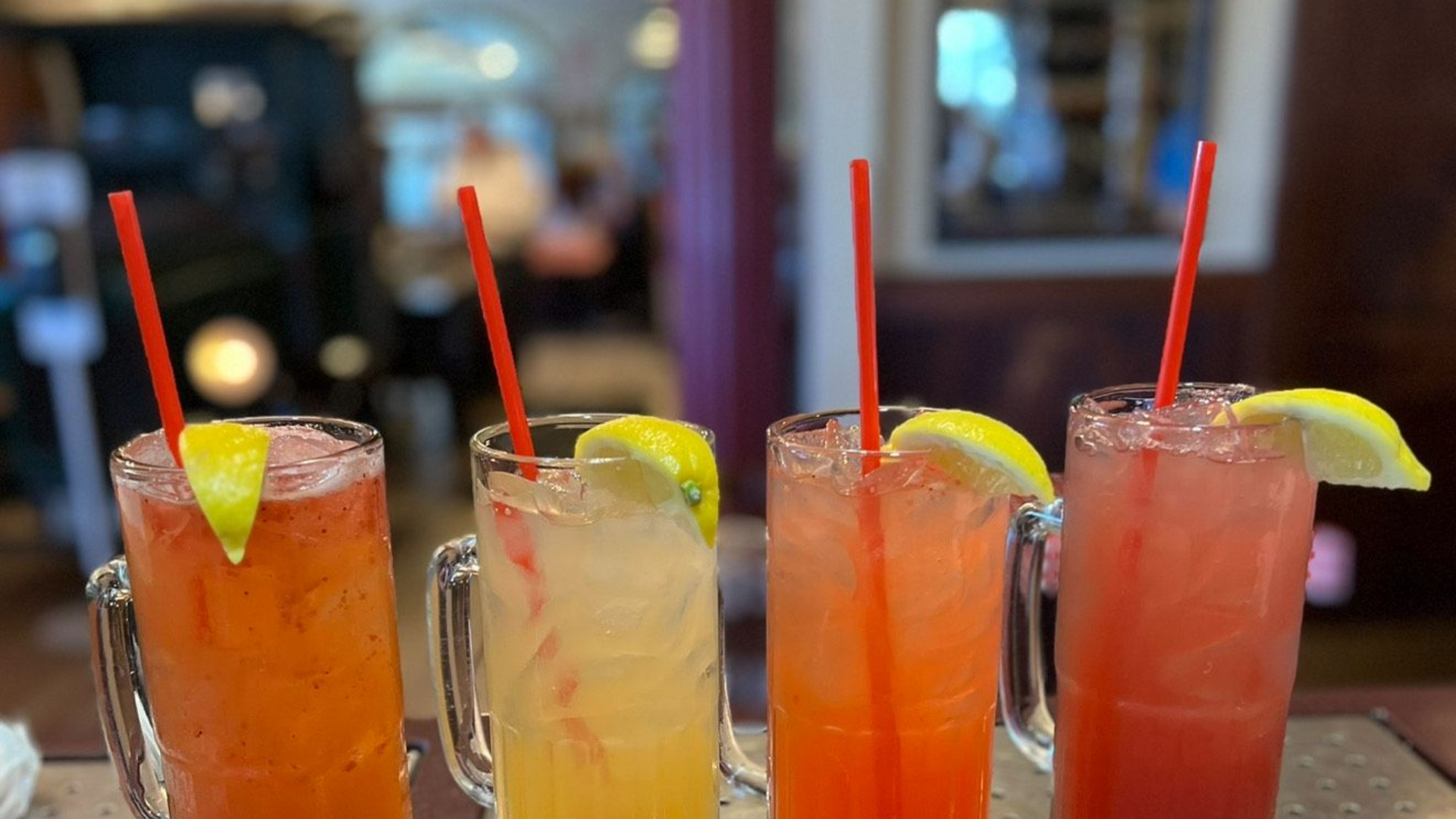 Four tall glasses of lemonade with red straws and lemon slices, on a bar.