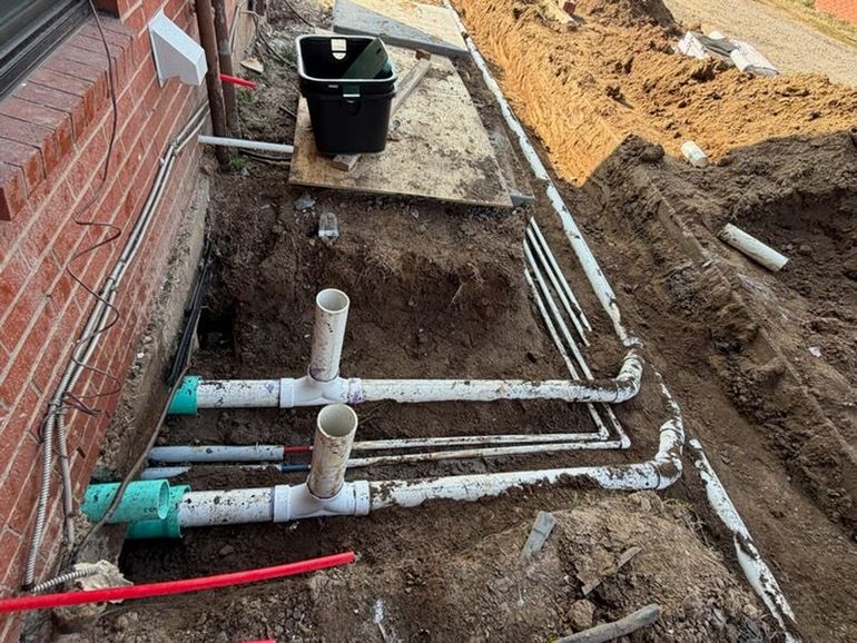 Trench beside brick wall with PVC pipes and utility lines being installed at a construction site
