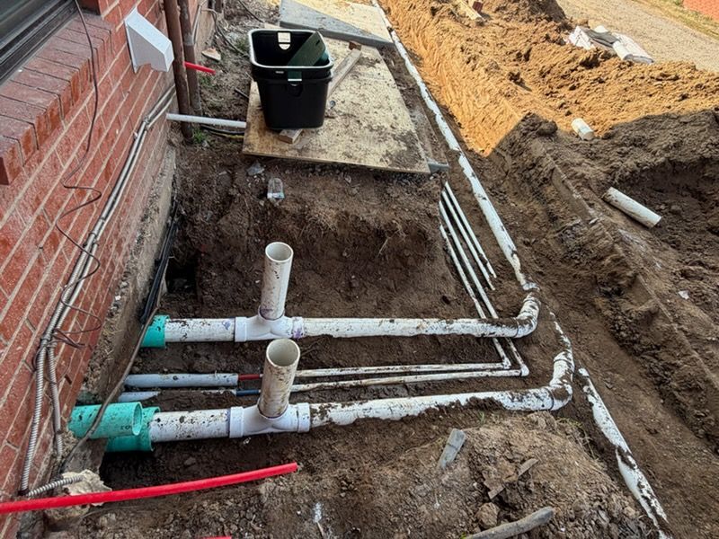 Trench beside brick wall with white PVC pipes and utility lines installed in muddy soil