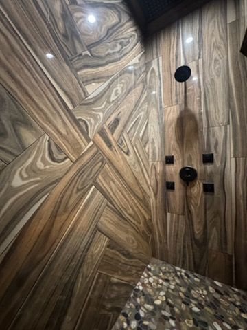 Wood-grain tiled shower with built-in niches, black fixtures, and pebble stone floor.