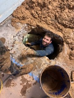 Man smiling in a muddy underground hole beside a large pipe during excavation work