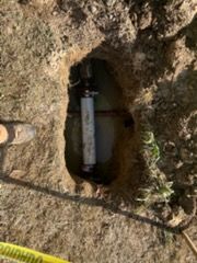 Small buried pipe or valve in a dirt hole beside yellow caution tape outdoors