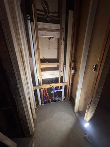 Narrow unfinished stairwell opening with exposed wood framing and pipes, viewed from above, lit by a small work light.