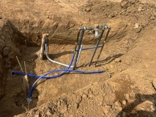 Water and utility lines exposed in a dirt trench with blue and black pipes and valves