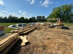 Construction site with lumber piles and excavator on a dirt foundation under a blue sky