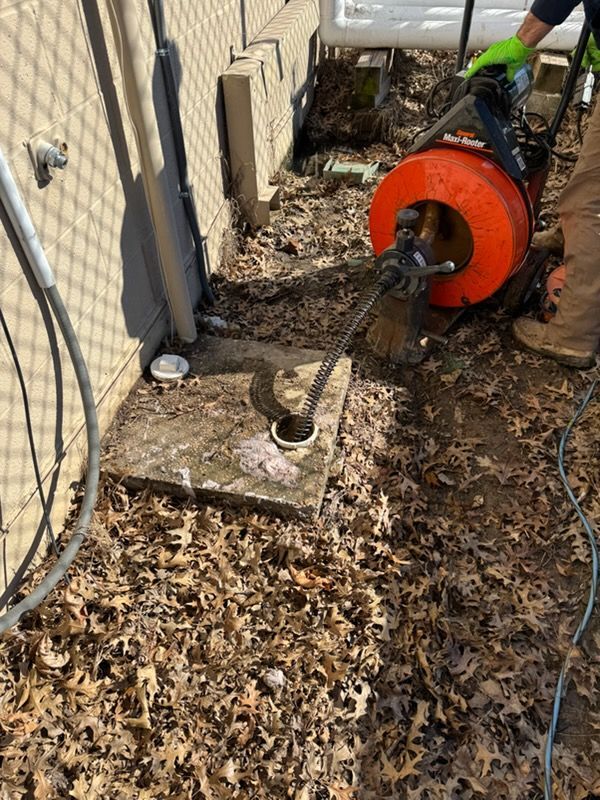 Worker using a drain auger to clear a clogged outdoor sewer line beside a house