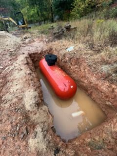 Red propane tank partially buried in muddy trench outdoors, with a black cap on top.