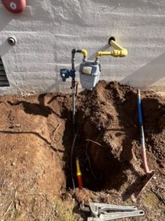 Exposed utility gas line and meter at a hole in the ground beside a house foundation, with tools nearby