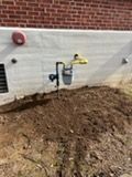 Yellow gas meter and pipe on house wall above a dirt pile by a brick foundation
