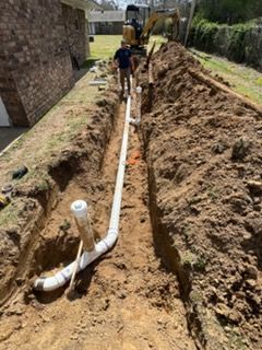 Worker by a trench installing white pipes beside a house, with an excavator in the background