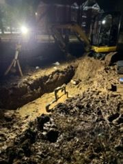 Nighttime excavation site with a backhoe digging a muddy trench under bright work lights.
