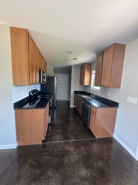 Narrow kitchen with wooden cabinets, black countertops, stainless appliances, and dark flooring.