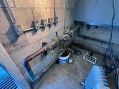 Utility room with exposed pipes, valves, bucket, and radiator against unfinished walls