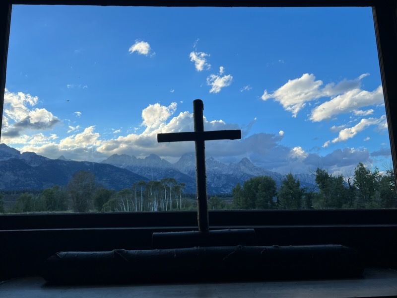 Silhouetted cross in a window, overlooking blue mountains and a bright sky with scattered clouds.