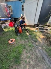 Person crouching beside a red pressure washer in a grassy yard near a shed