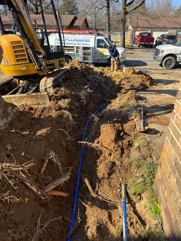 Excavator digging a trench beside a brick building, with workers and utility trucks on a muddy construction site.