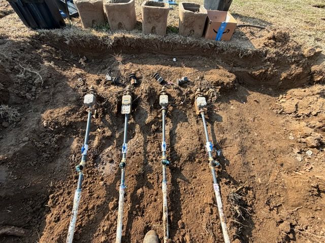Four irrigation pipes laid in a trench beside a brick wall on a dirt construction site