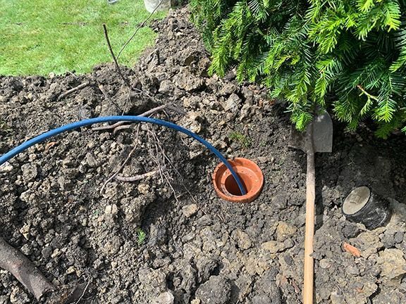 Blue hose watering a small orange pot in freshly dug soil beside a green shrub