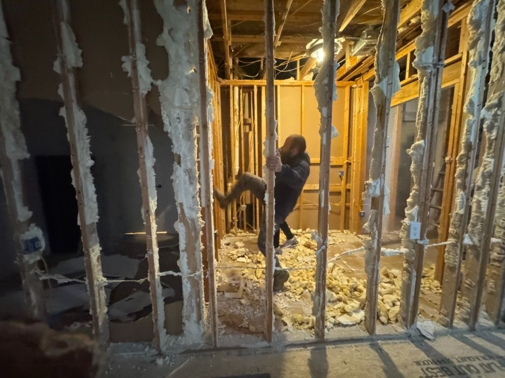 Worker in a gutted room with exposed studs and insulation, likely under renovation.
