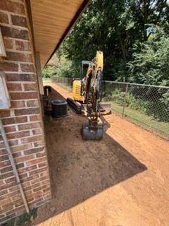 Mini excavator on dirt beside a brick house, digging a trench along a chain-link fence.